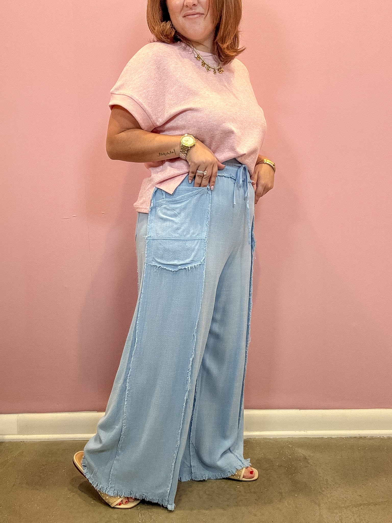 A woman modeling a light pink short-sleeve t-shirt and light blue wide-leg denim pants with raw hems, standing against a solid pink wall.