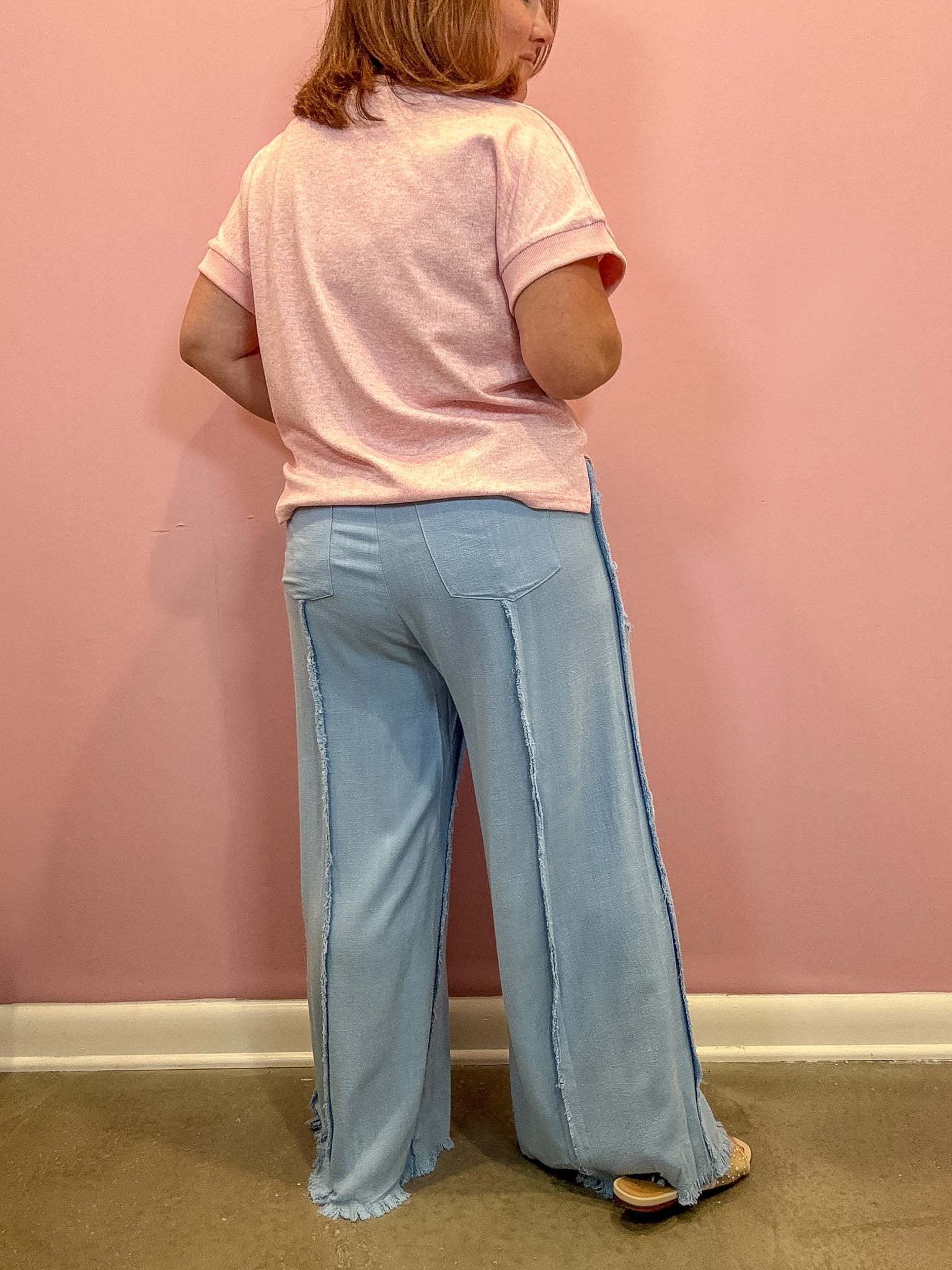 A person shown from the back wearing a light pink short-sleeve t-shirt and light blue wide-leg denim pants with side seam details, standing against a pink wall.