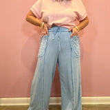 A woman modeling a light pink short-sleeve knit top and light blue wide-leg denim pants with fringe and patch pockets, standing against a pink wall.