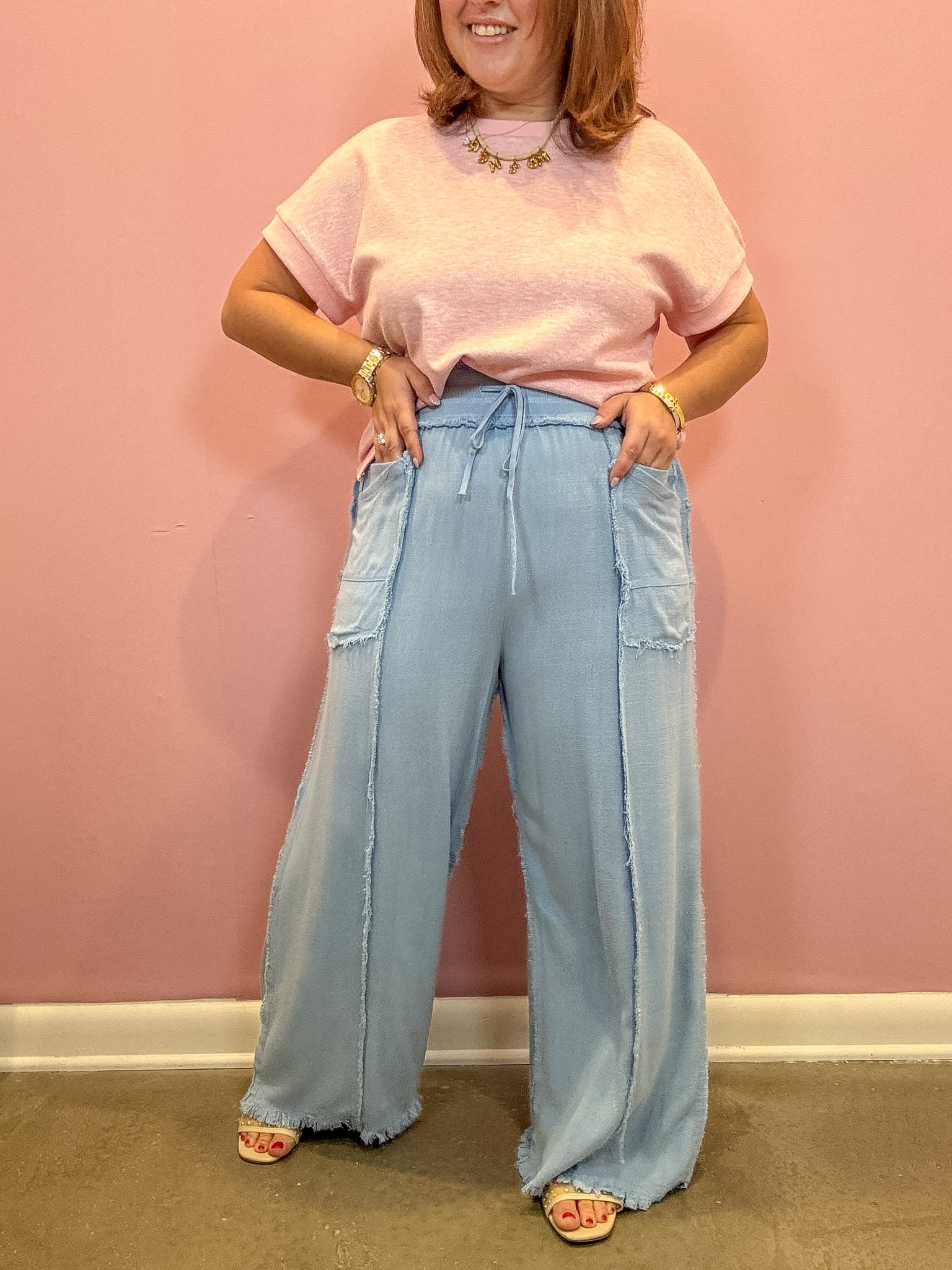 A woman modeling a light pink short-sleeve knit top and light blue wide-leg denim pants with fringe and patch pockets, standing against a pink wall.