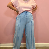 A woman wearing a light pink short-sleeve t-shirt and light blue wide-leg drawstring pants, standing against a pink wall.