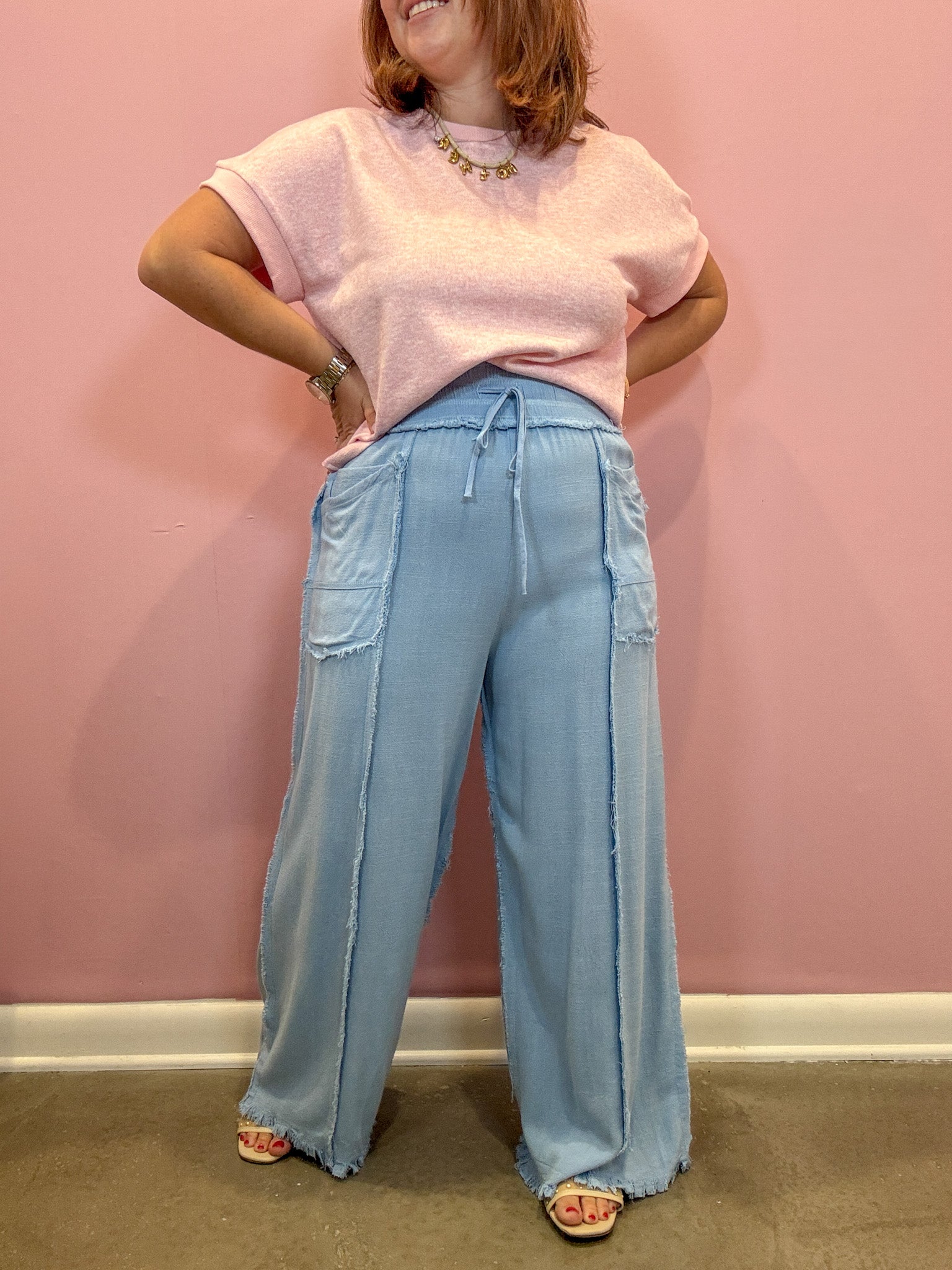 A woman wearing a light pink short-sleeve t-shirt and light blue wide-leg drawstring pants, standing against a pink wall.