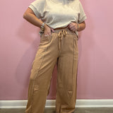 A person modeling wide-leg tan pants with a drawstring waist and a light beige short-sleeve top, standing against a pink wall.