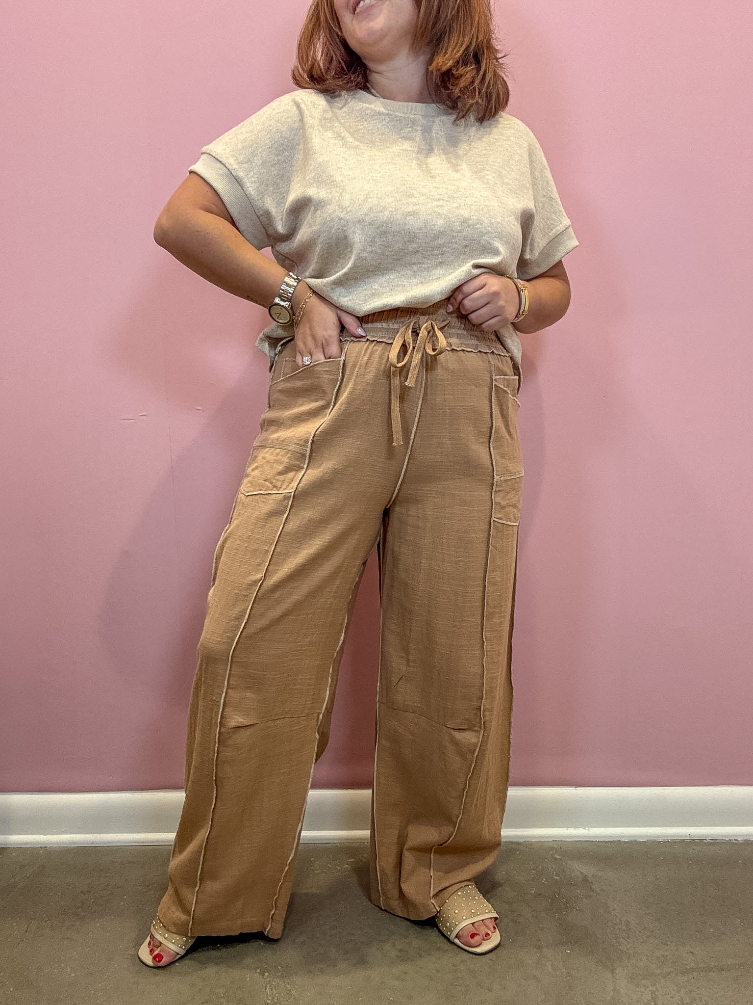 A person modeling wide-leg tan pants with a drawstring waist and a light beige short-sleeve top, standing against a pink wall.