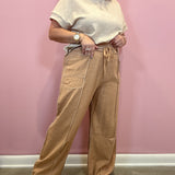 A woman modeling a pair of wide-leg tan pants with a drawstring waist and contrast stitching, paired with a cream knit top and beige sandals, standing against a solid pink wall.