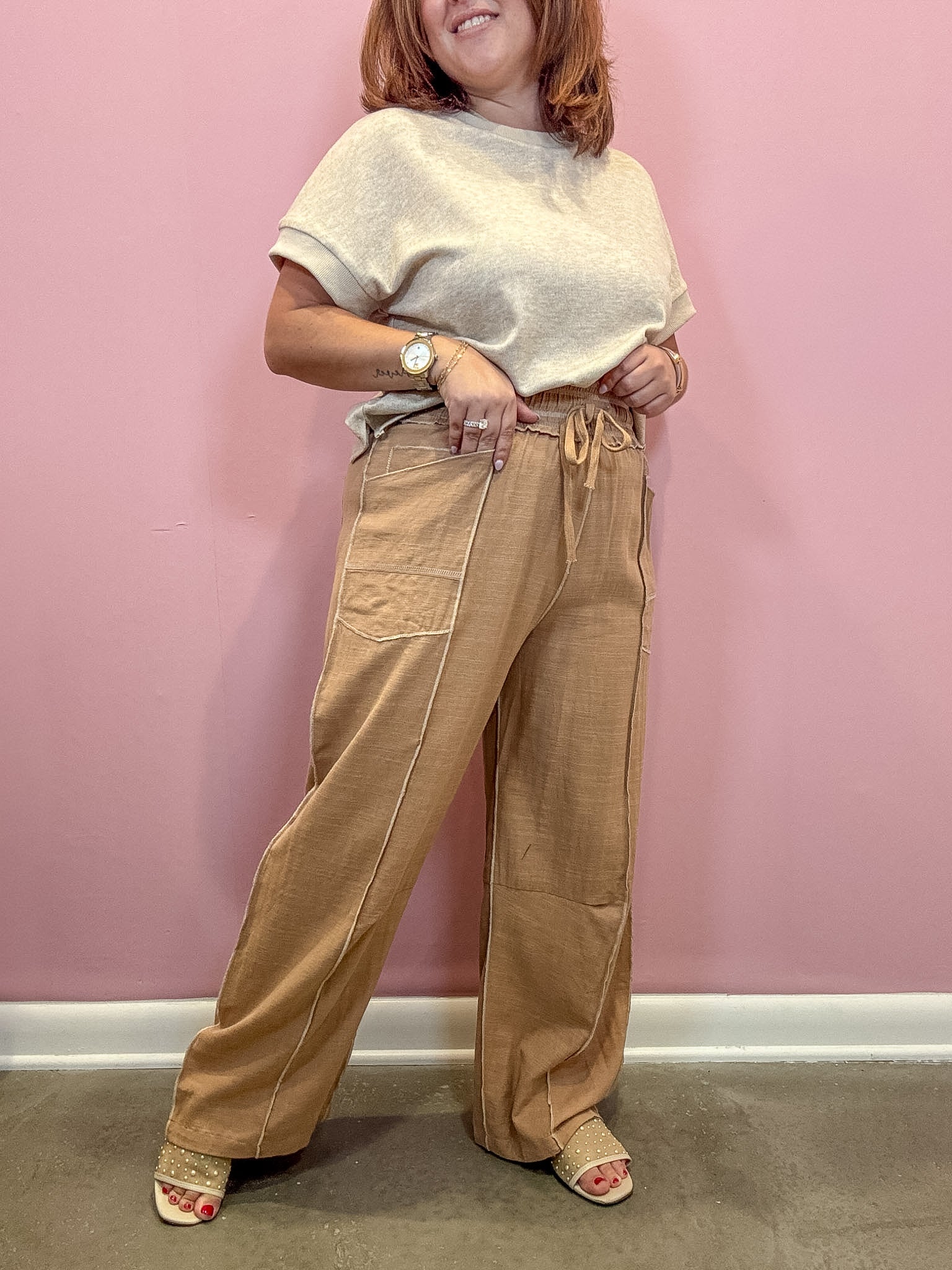 A woman modeling a pair of wide-leg tan pants with a drawstring waist and contrast stitching, paired with a cream knit top and beige sandals, standing against a solid pink wall.