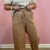 A person wearing loose, wide-leg tan pants with a drawstring waist and a light beige short-sleeve top, standing against a pink wall.