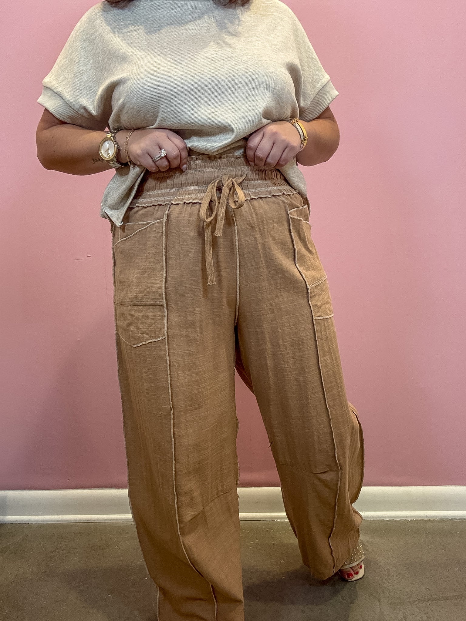 A person wearing loose, wide-leg tan pants with a drawstring waist and a light beige short-sleeve top, standing against a pink wall.