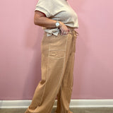 A woman modeling a pair of wide-leg tan pants with contrast white stitching, paired with a light beige t-shirt, standing against a solid pink wall.
