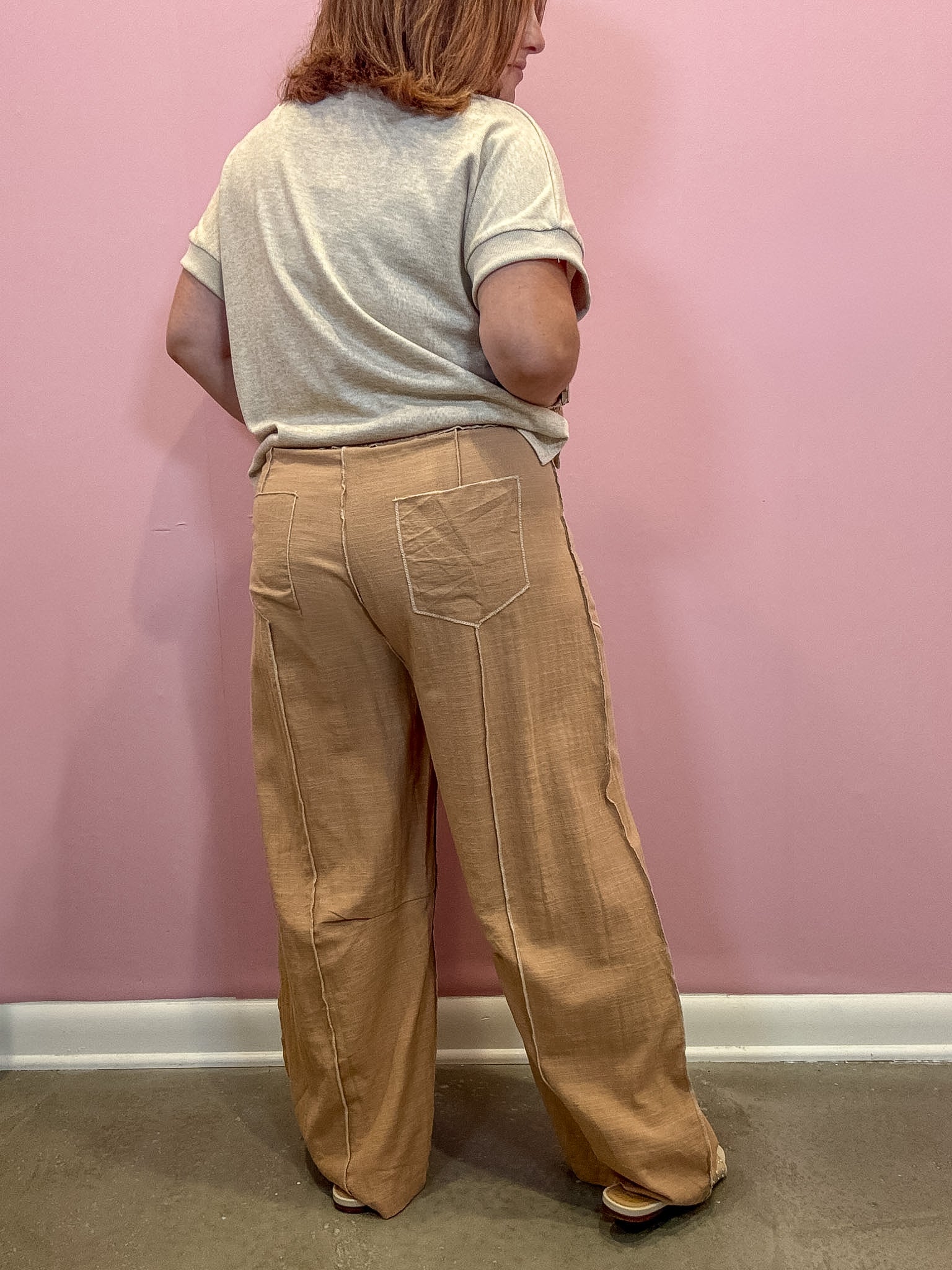 A person shown from the back wearing tan wide-leg linen trousers and a light gray t-shirt, standing against a pink wall.