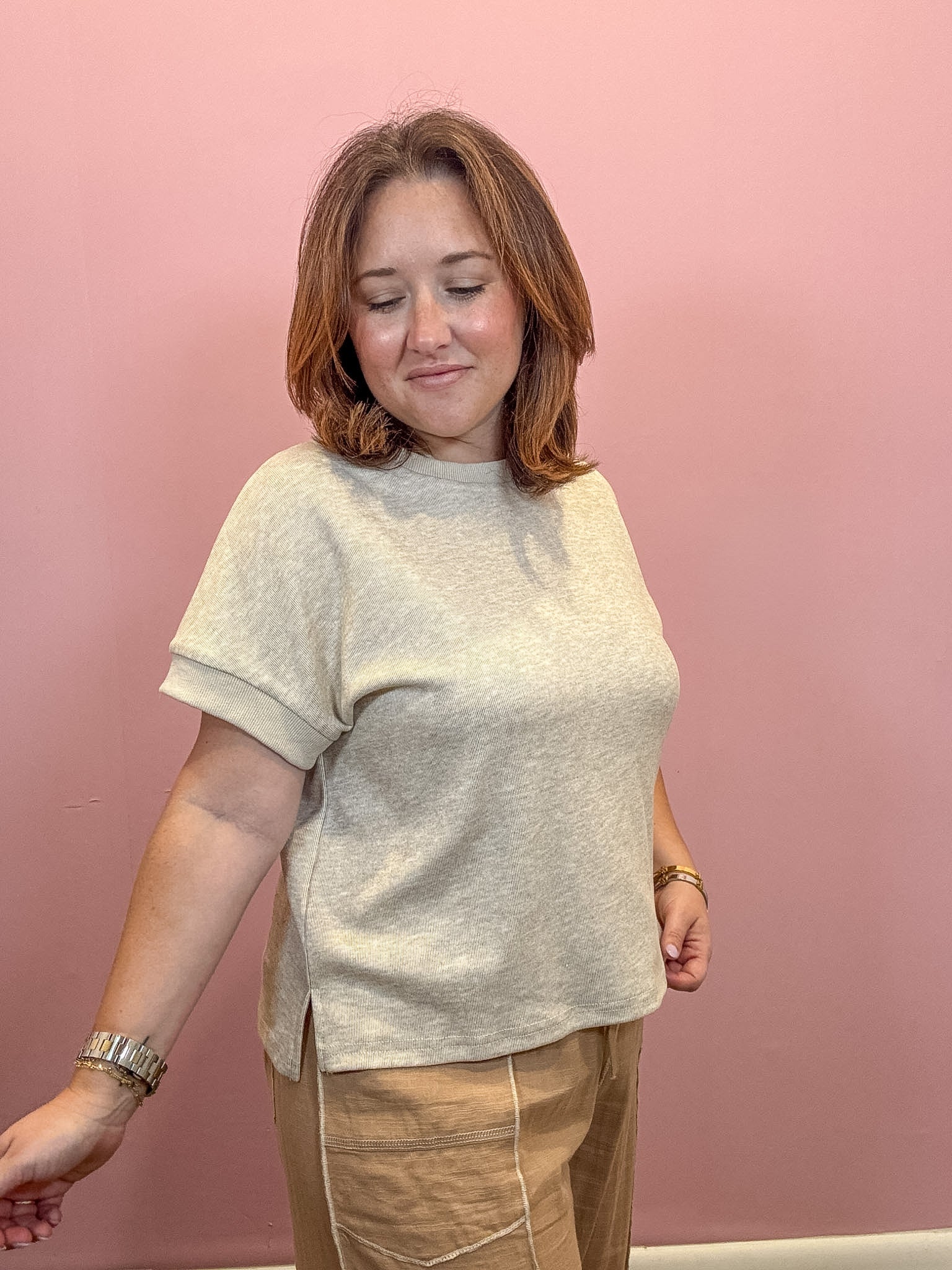 A woman with shoulder-length brown hair wearing a light beige short-sleeve knit top and tan pants, standing against a solid dusty pink wall.