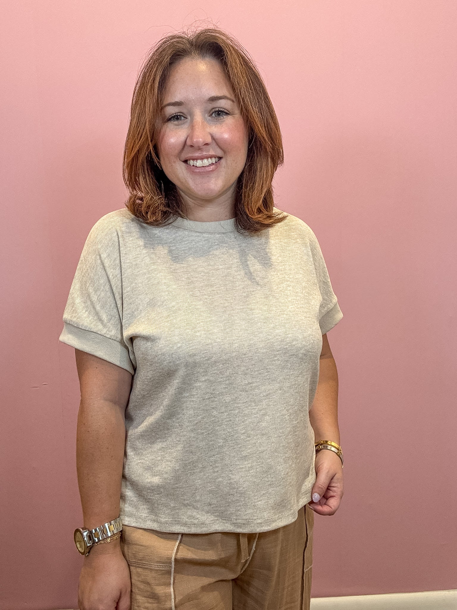 A smiling woman with shoulder-length brown hair wearing a light beige short-sleeve ribbed t-shirt and tan pants, standing against a solid pink wall.