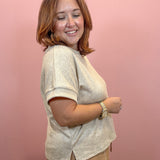 A woman with shoulder-length auburn hair is shown from the side, wearing a light beige short-sleeve knit top and tan pants against a solid pink wall.
