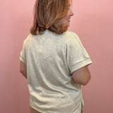 A woman with shoulder-length brown hair is shown from the back wearing a light beige heathered short-sleeve t-shirt and tan pants, standing against a solid pink wall.