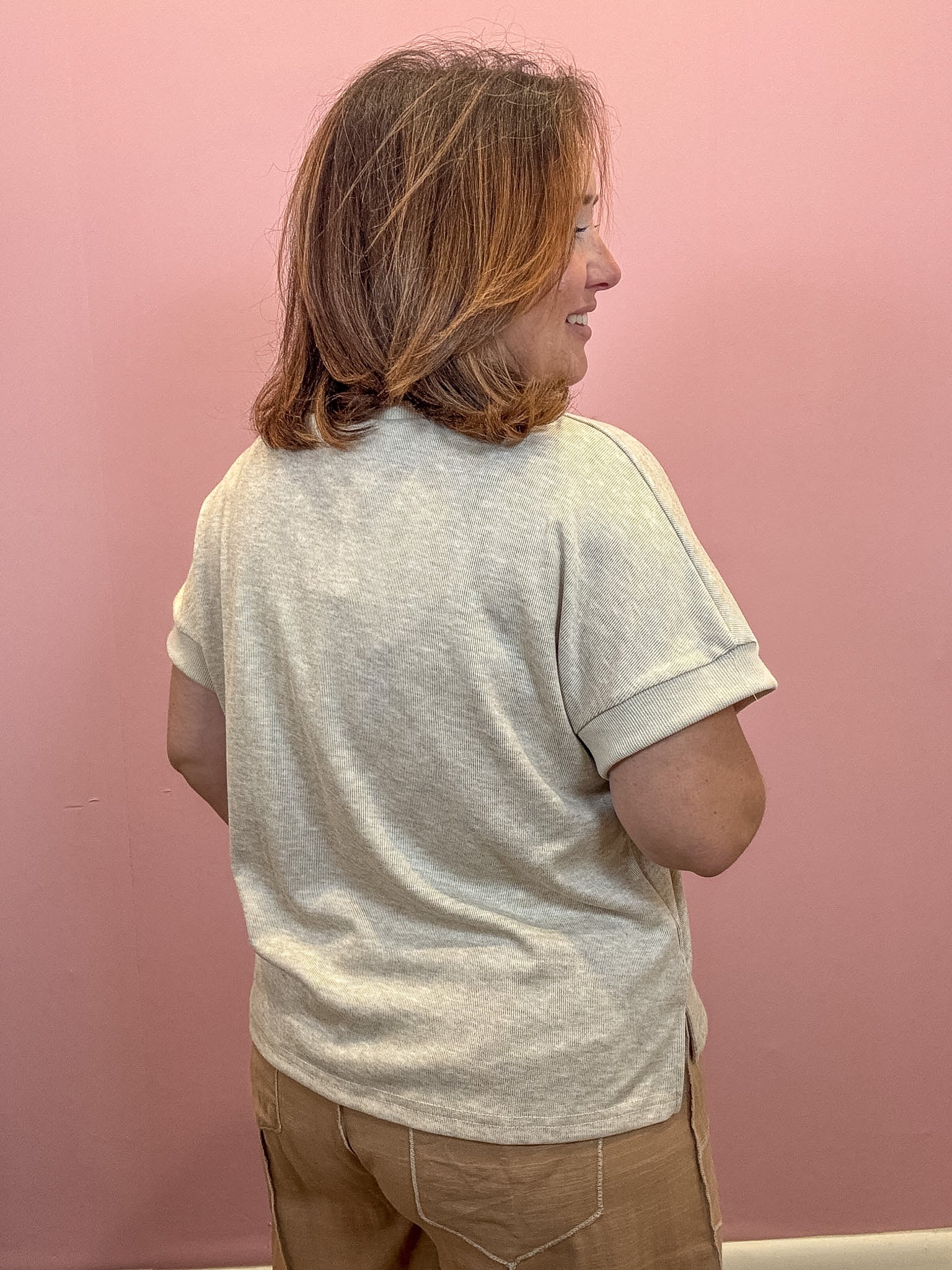 A woman with shoulder-length brown hair is shown from the back wearing a light beige heathered short-sleeve t-shirt and tan pants, standing against a solid pink wall.