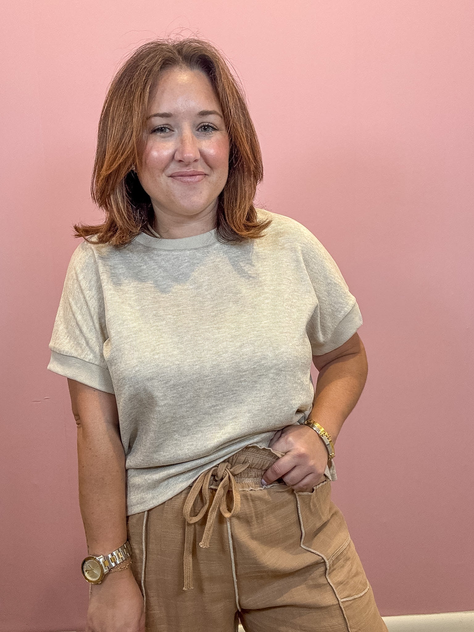 A woman with shoulder-length brown hair stands against a solid pink wall, wearing a light beige heathered short-sleeve t-shirt and tan drawstring pants.