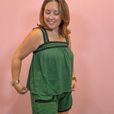 A woman modeling a green two-piece lounge set consisting of a sleeveless top and matching shorts, both featuring black braided trim details, posed against a solid pink wall.