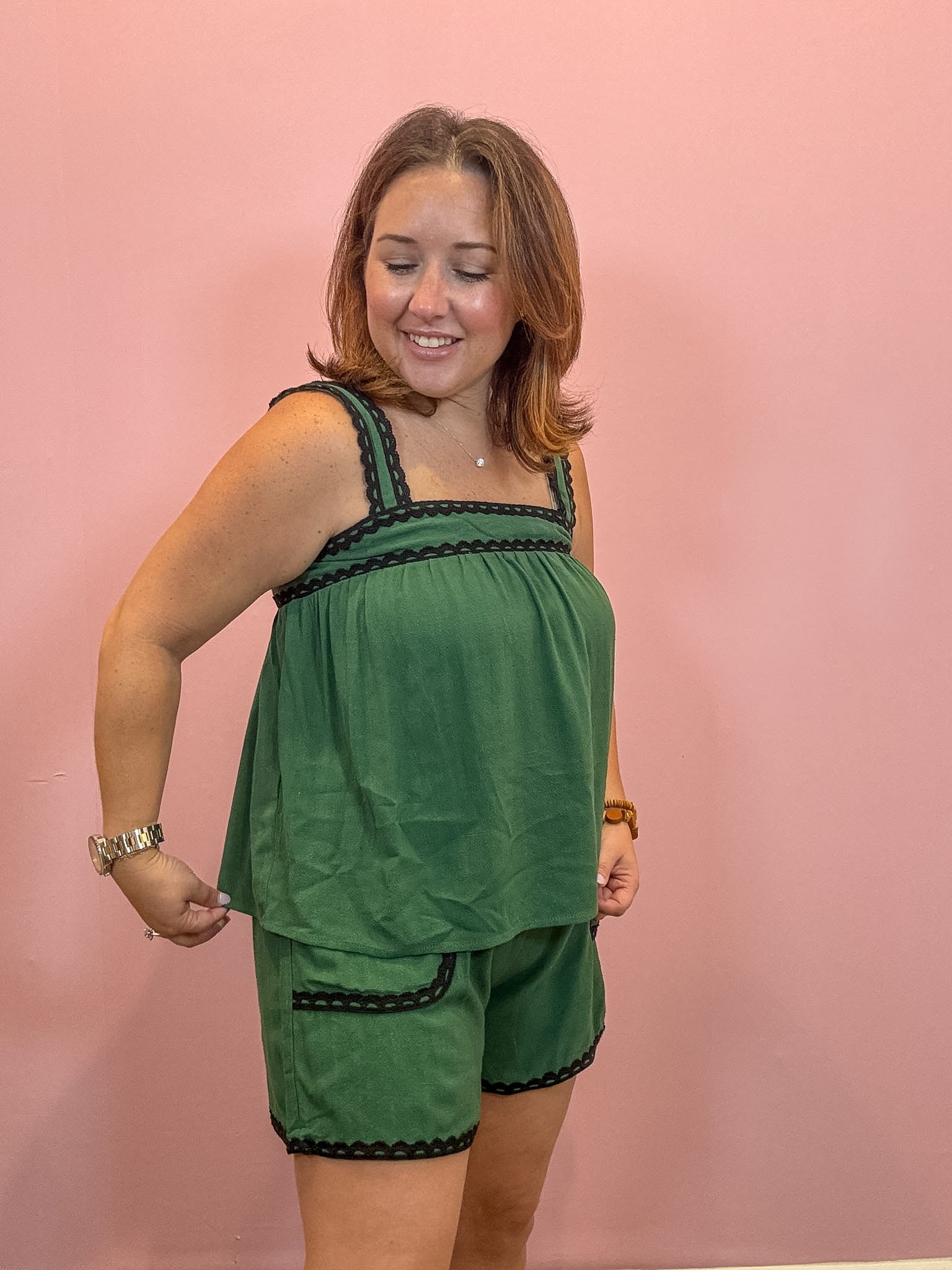 A woman modeling a green two-piece lounge set consisting of a sleeveless top and matching shorts, both featuring black braided trim details, posed against a solid pink wall.