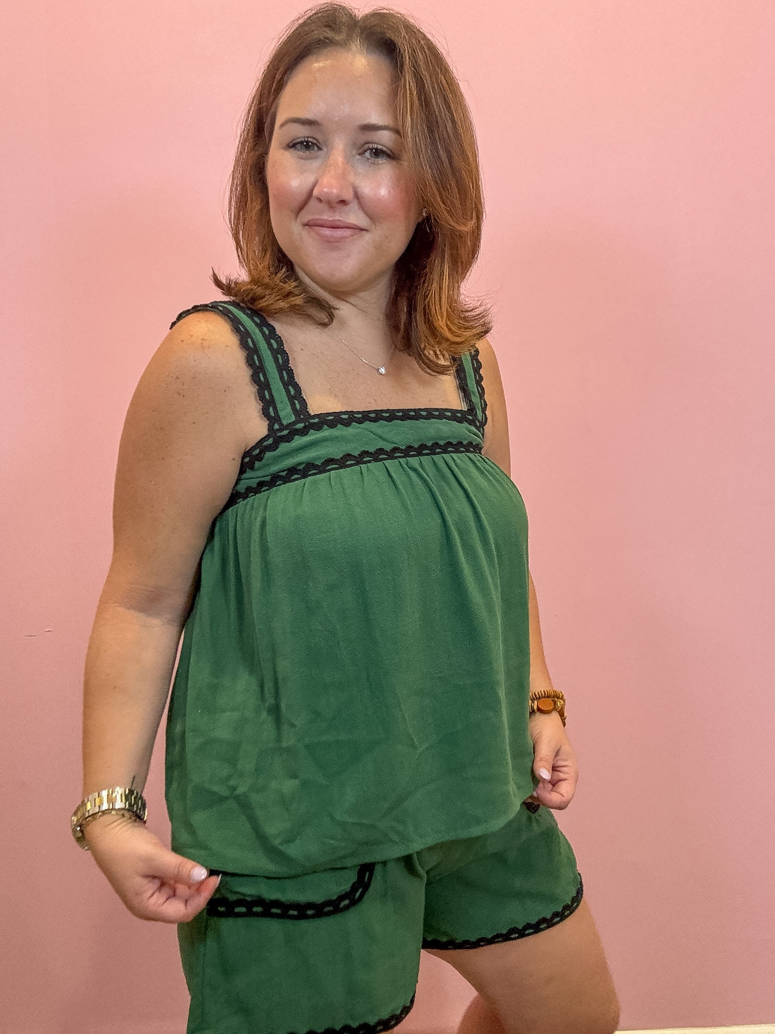 A woman modeling a two-piece green lounge set with black lace trim, consisting of a sleeveless top and matching shorts, set against a solid pink background.