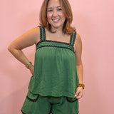 A woman modeling a green two-piece lounge set with black scalloped trim, consisting of a sleeveless top and matching shorts, posed against a solid pink wall.