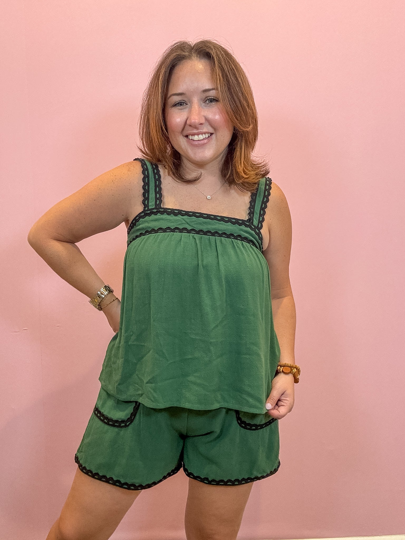 A woman modeling a green two-piece lounge set with black scalloped trim, consisting of a sleeveless top and matching shorts, posed against a solid pink wall.
