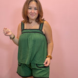 A woman modeling a green two-piece lounge set with black trim, consisting of a sleeveless top and matching shorts, set against a solid pink background.