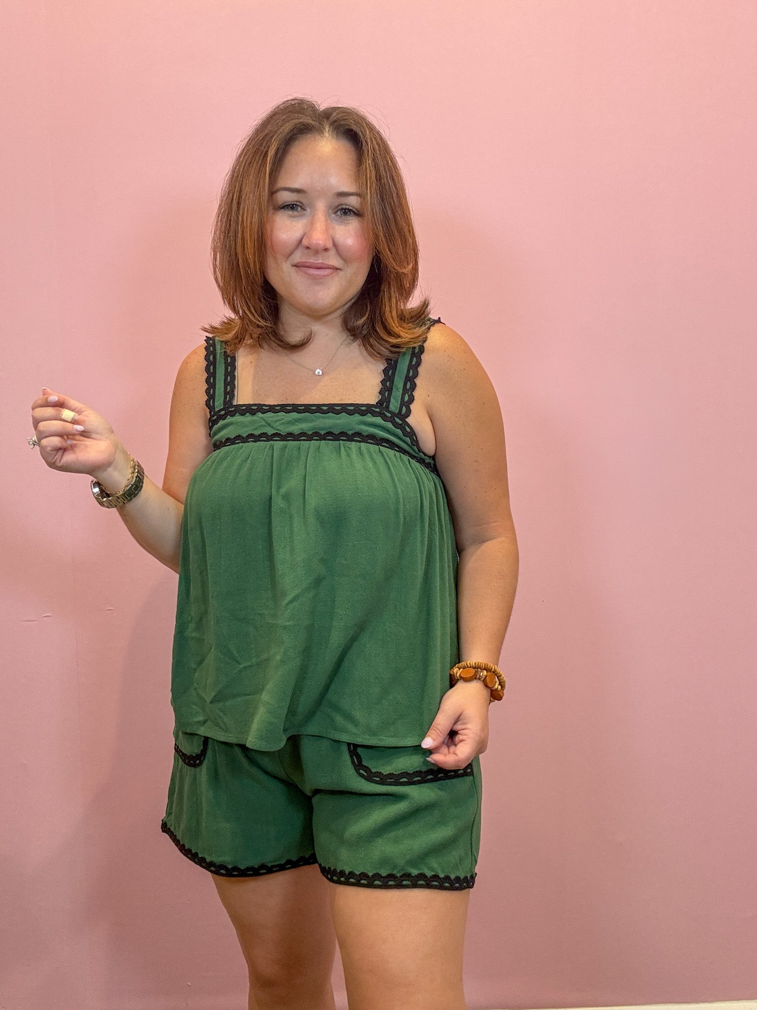 A woman modeling a green two-piece lounge set with black trim, consisting of a sleeveless top and matching shorts, set against a solid pink background.