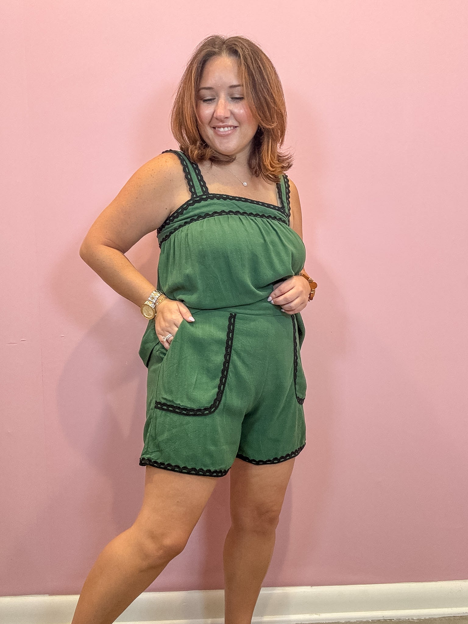 A woman modeling a green sleeveless romper with black trim, standing against a solid pink wall.