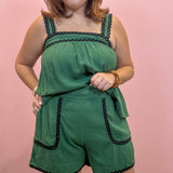 A woman modeling a green sleeveless romper with black lace trim, standing against a solid pink wall.