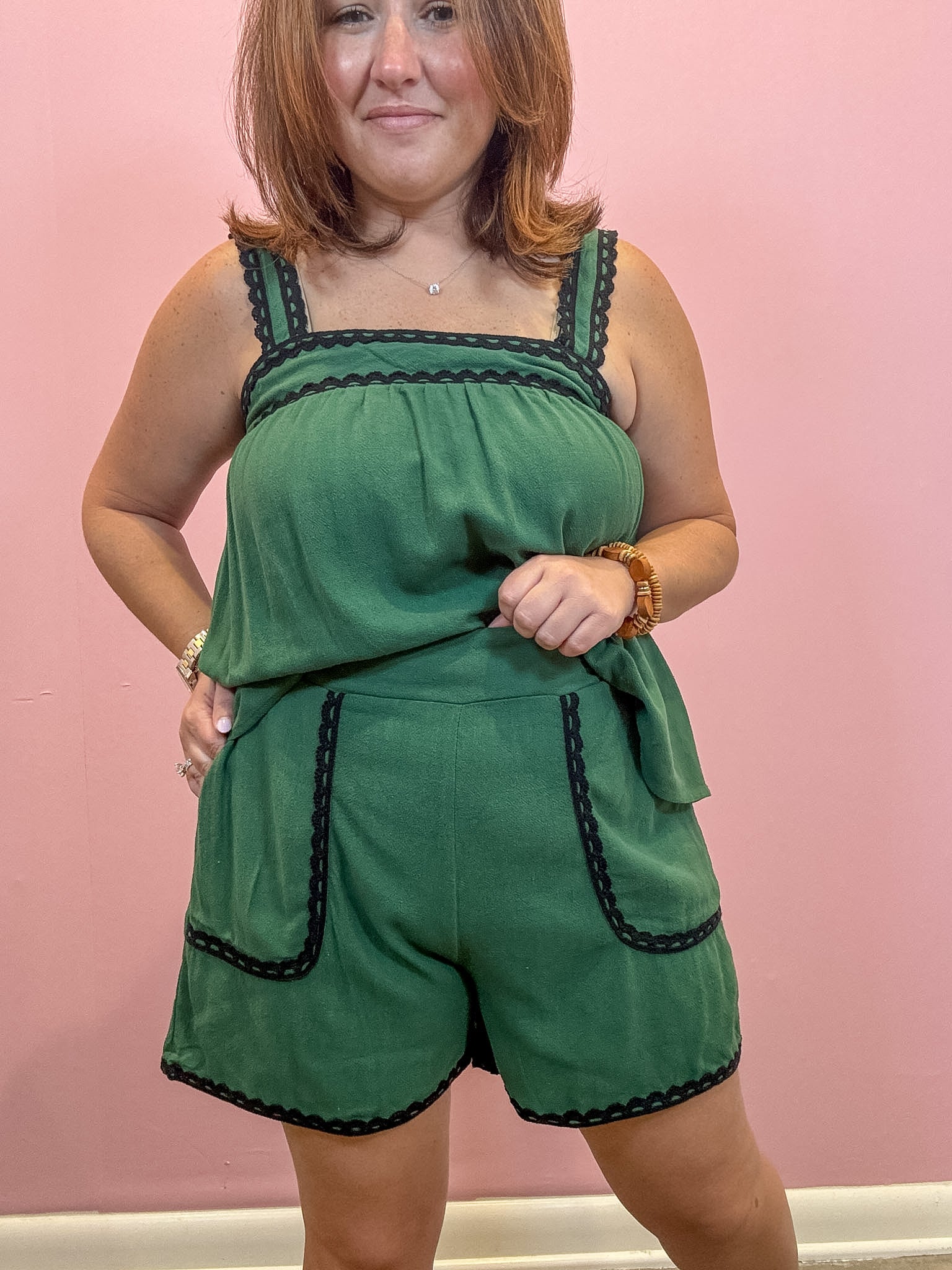 A woman modeling a green sleeveless romper with black lace trim, standing against a solid pink wall.