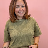 A woman with shoulder-length brown hair smiling, wearing a heathered olive green short-sleeve t-shirt with a chest pocket and beige pants, standing against a solid pink background.
