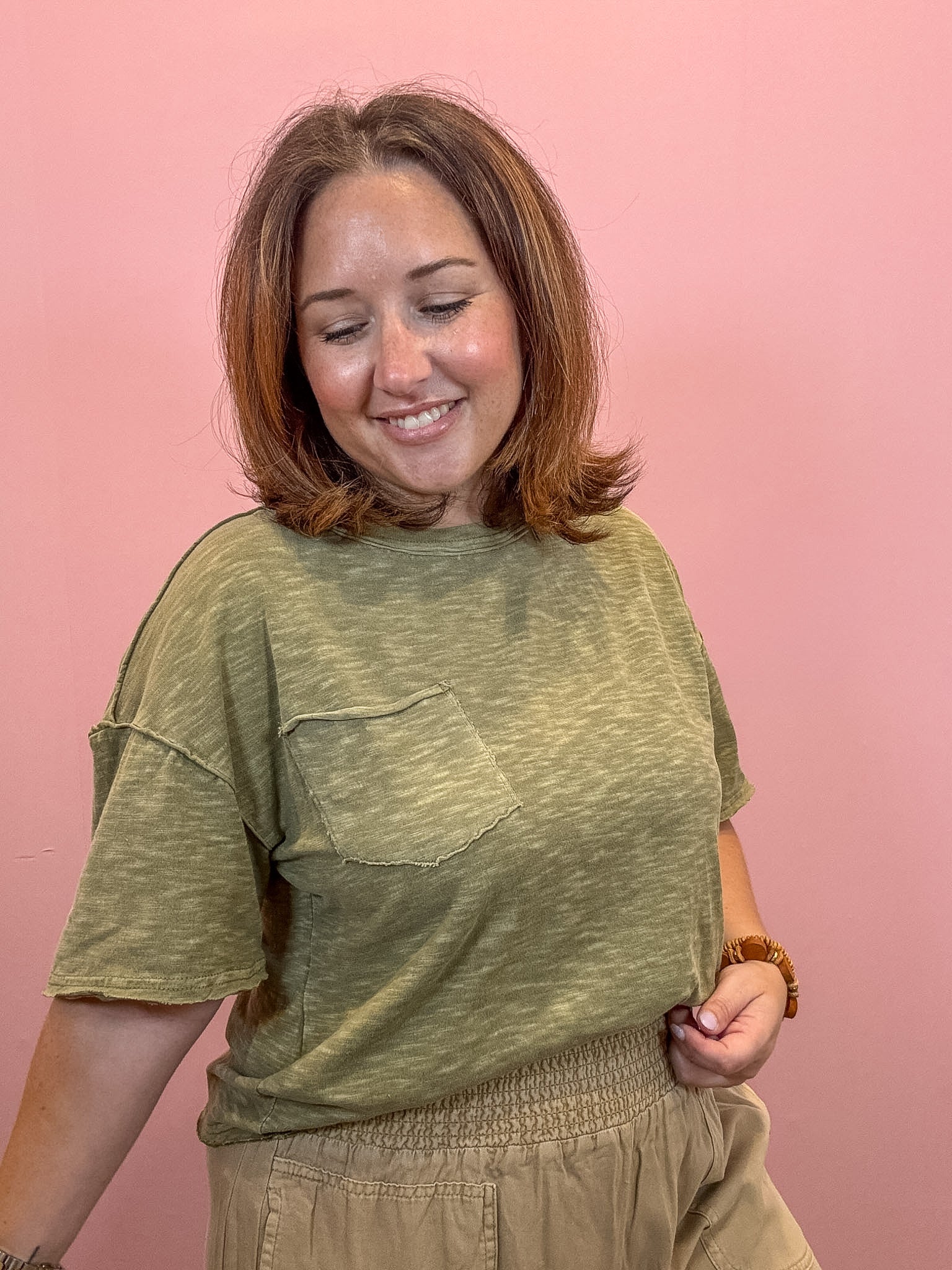 A woman with shoulder-length brown hair smiling, wearing a heathered olive green short-sleeve t-shirt with a chest pocket and beige pants, standing against a solid pink background.