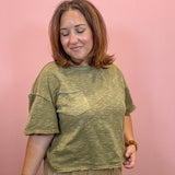 A woman with shoulder-length brown hair smiling, wearing a heathered olive green short-sleeve t-shirt with a chest pocket and beige pants, standing against a solid pink background.