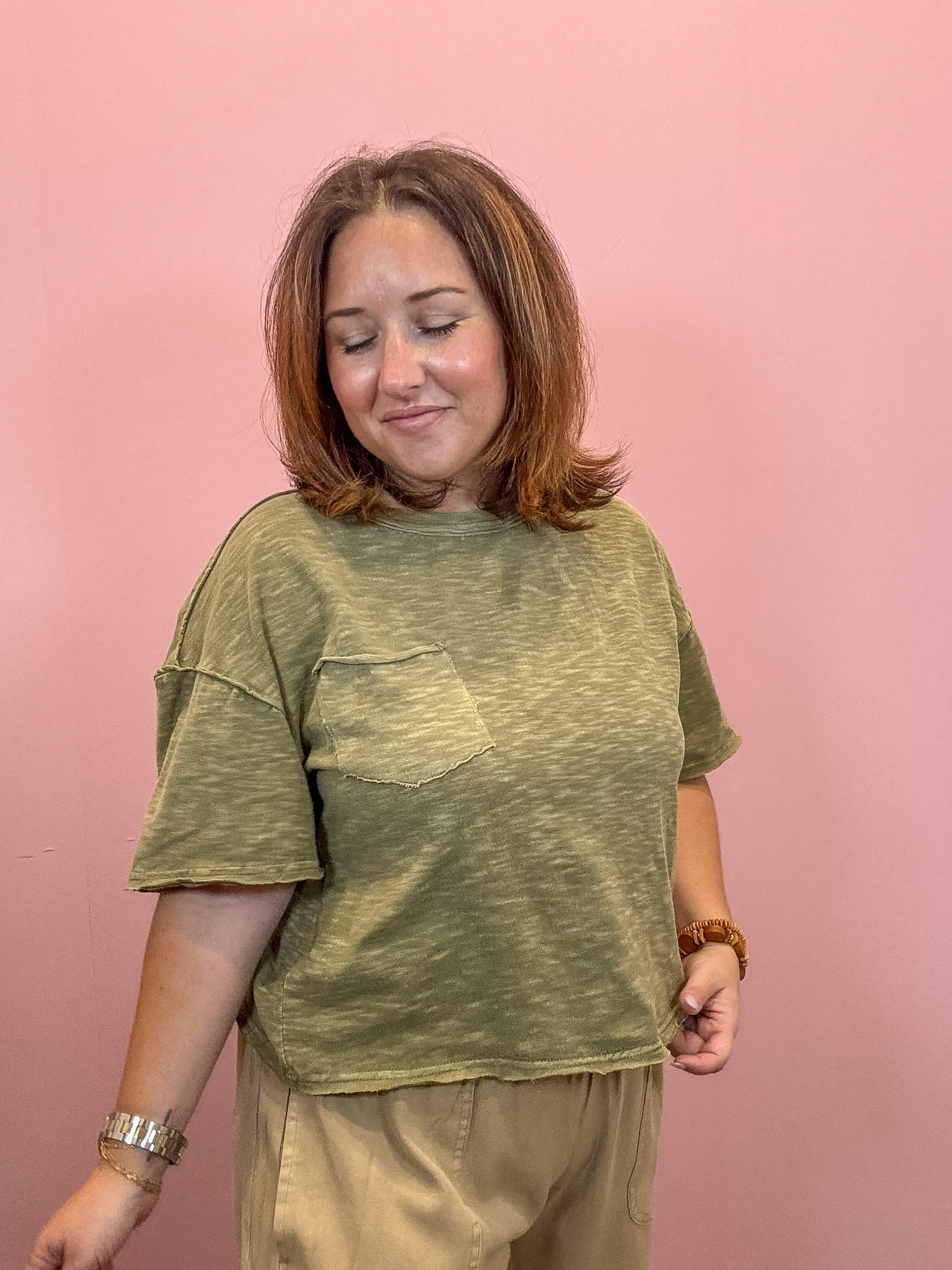 A woman with shoulder-length brown hair smiling, wearing a heathered olive green short-sleeve t-shirt with a chest pocket and beige pants, standing against a solid pink background.