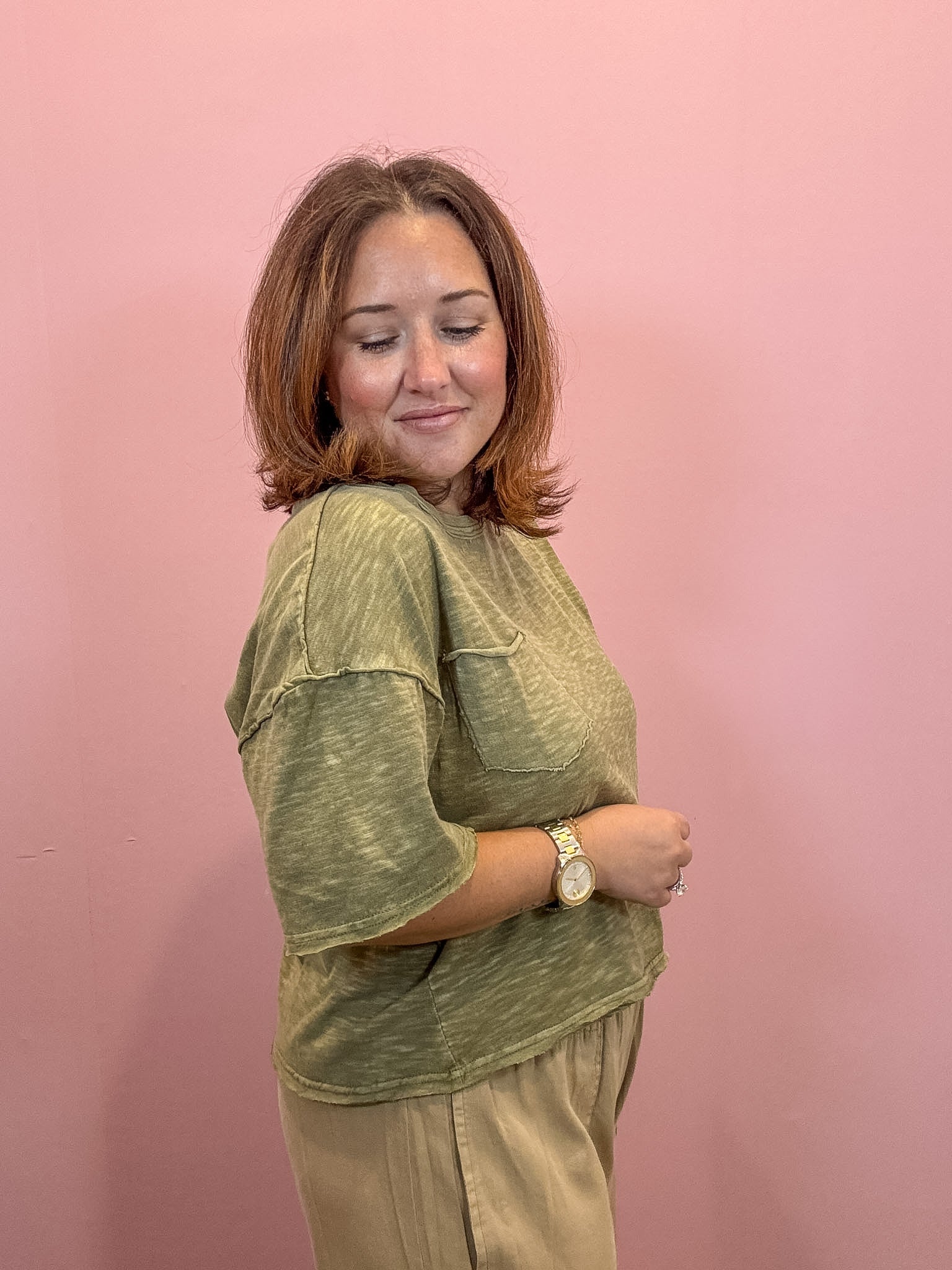 A woman with shoulder-length brown hair smiling, wearing a heathered olive green short-sleeve t-shirt with a chest pocket and beige pants, standing against a solid pink background.