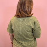 A woman with shoulder-length brown hair smiling, wearing a heathered olive green short-sleeve t-shirt with a chest pocket and beige pants, standing against a solid pink background.