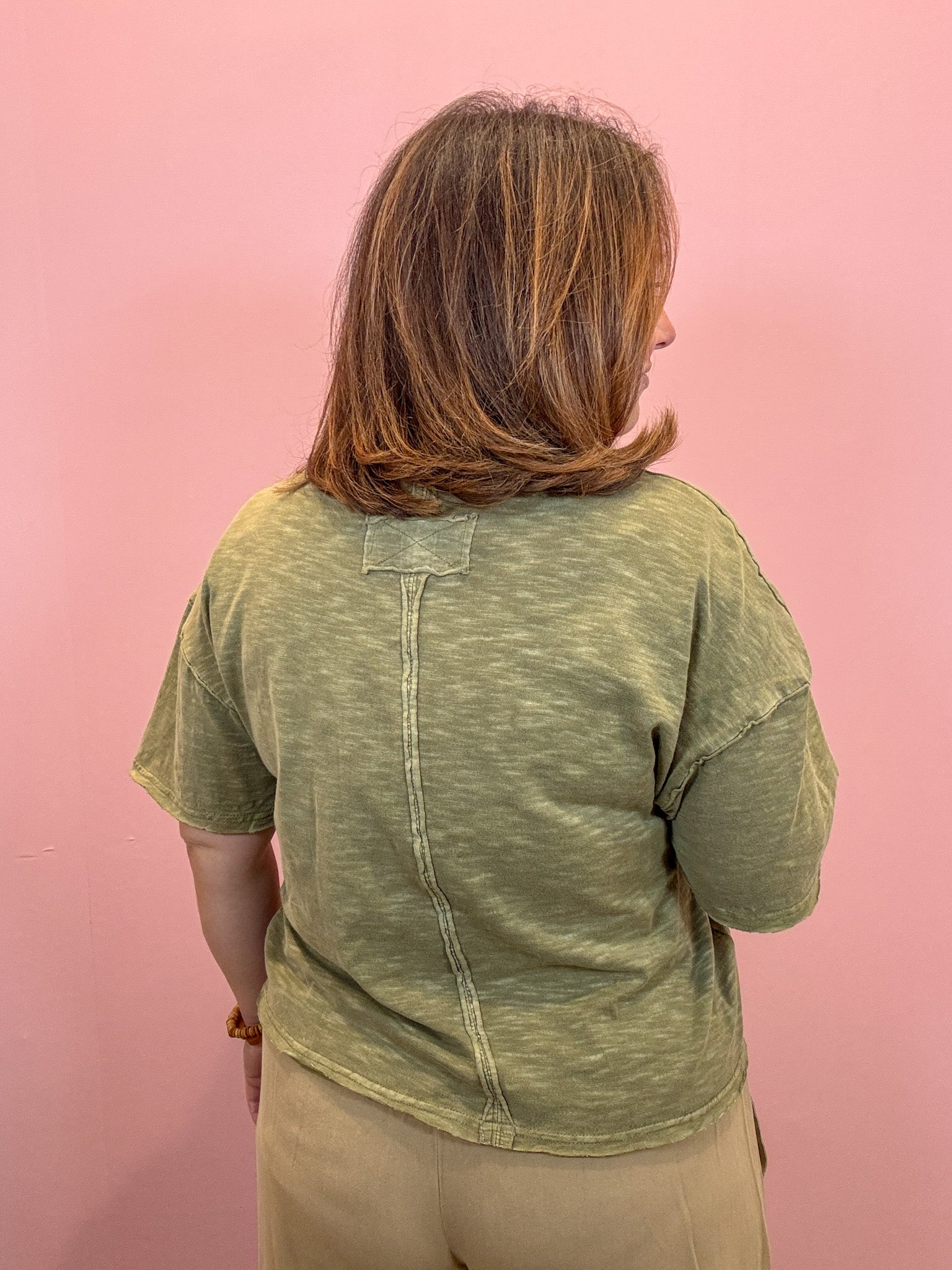 A woman with shoulder-length brown hair smiling, wearing a heathered olive green short-sleeve t-shirt with a chest pocket and beige pants, standing against a solid pink background.