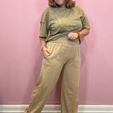A woman modeling a two-piece outfit consisting of a short-sleeve olive green t-shirt and high-waisted beige wide-leg pants, standing against a solid pink wall.