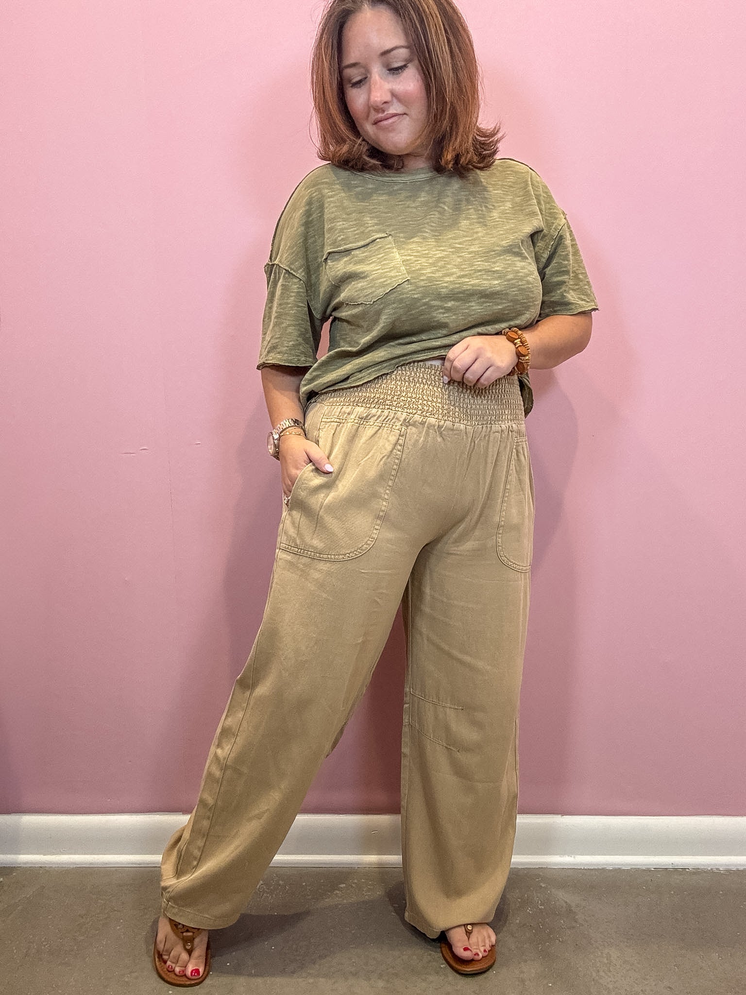 A woman modeling a two-piece outfit consisting of a short-sleeve olive green t-shirt and high-waisted beige wide-leg pants, standing against a solid pink wall.