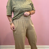 A woman modeling a two-piece outfit consisting of a heathered olive green short-sleeve t-shirt and beige wide-leg pants with a smocked waist, standing against a pink wall.
