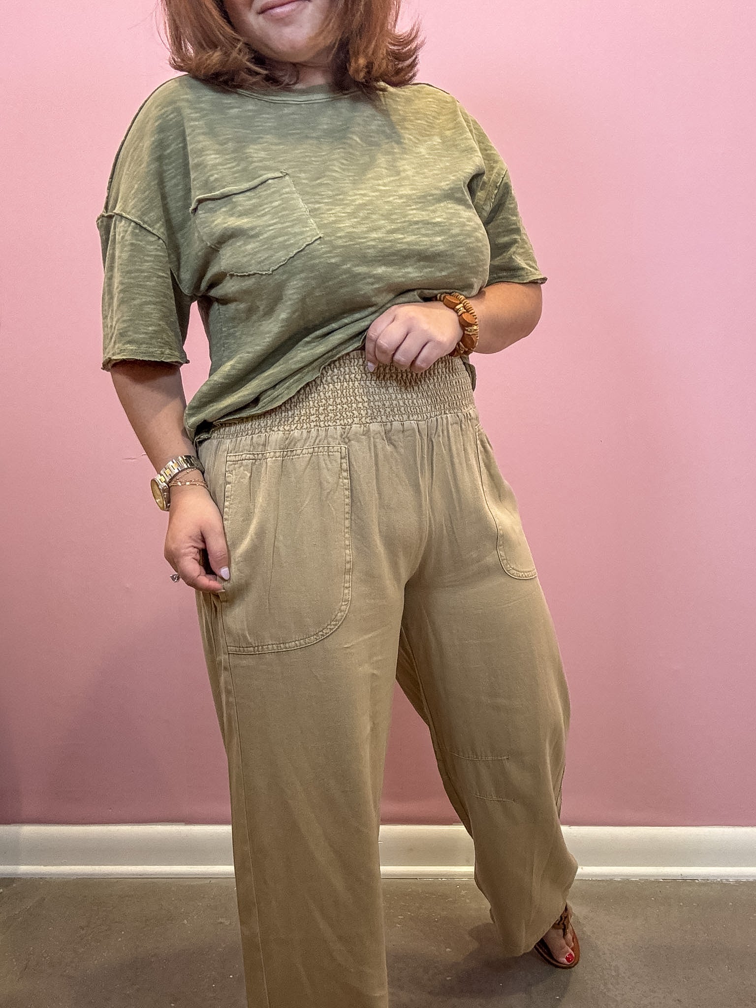 A woman modeling a two-piece outfit consisting of a heathered olive green short-sleeve t-shirt and beige wide-leg pants with a smocked waist, standing against a pink wall.