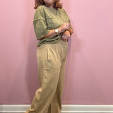 A woman modeling a two-piece outfit consisting of a sage green short-sleeve top and beige wide-leg pants, standing against a solid pink wall.
