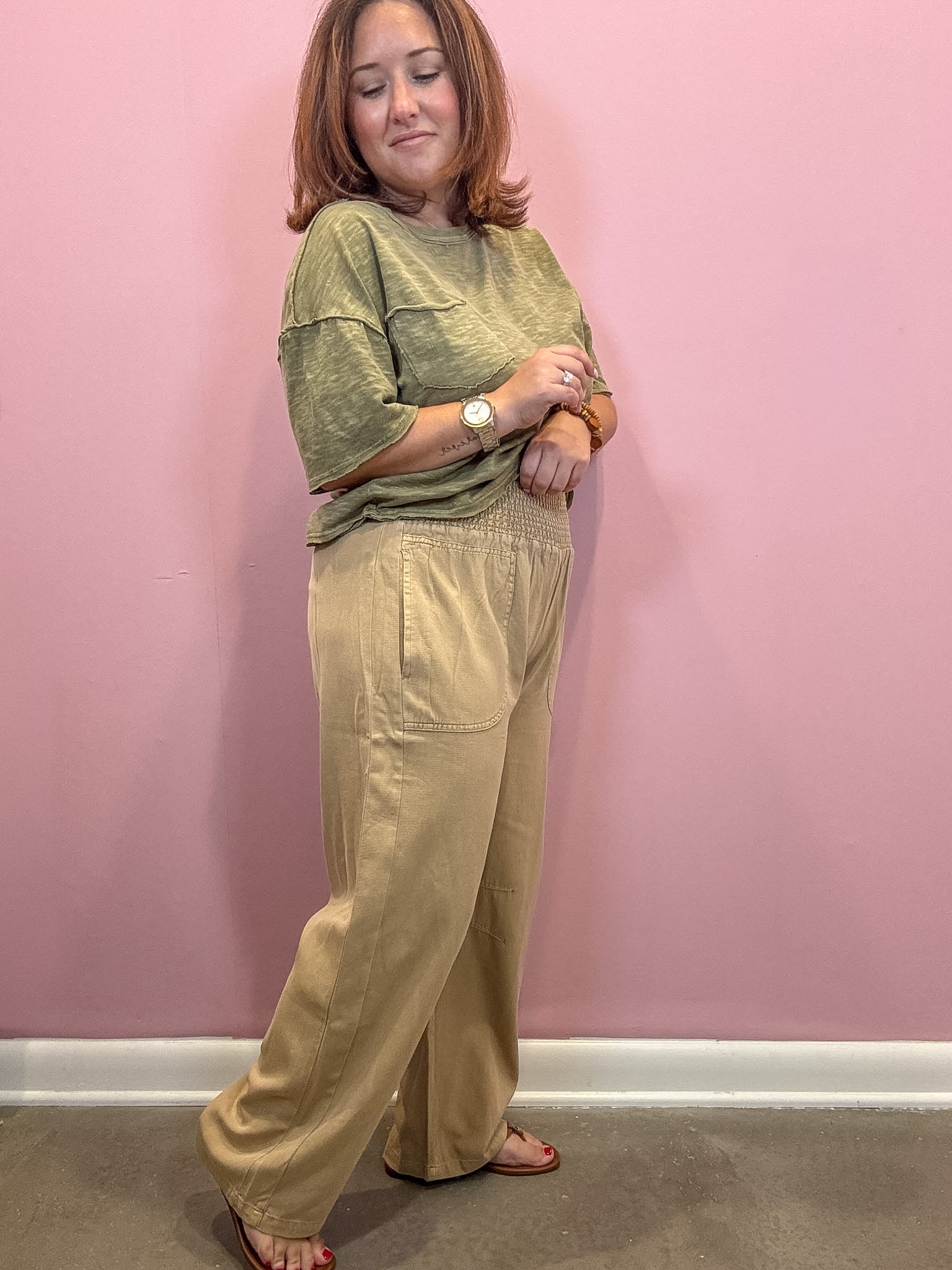 A woman modeling a two-piece outfit consisting of a sage green short-sleeve top and beige wide-leg pants, standing against a solid pink wall.