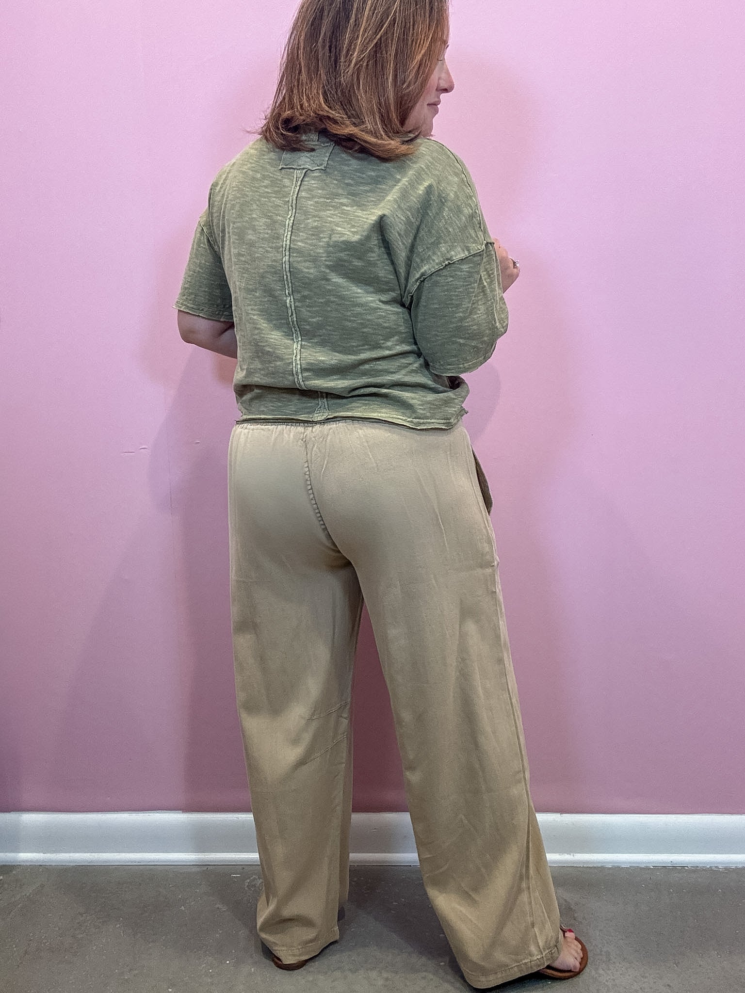 A woman shown from the back wearing a heathered sage green short-sleeve top and beige wide-leg pants, standing against a solid pink wall.