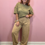 A woman modeling a two-piece outfit consisting of a short-sleeve olive green top and matching beige wide-leg pants, standing against a pink wall.