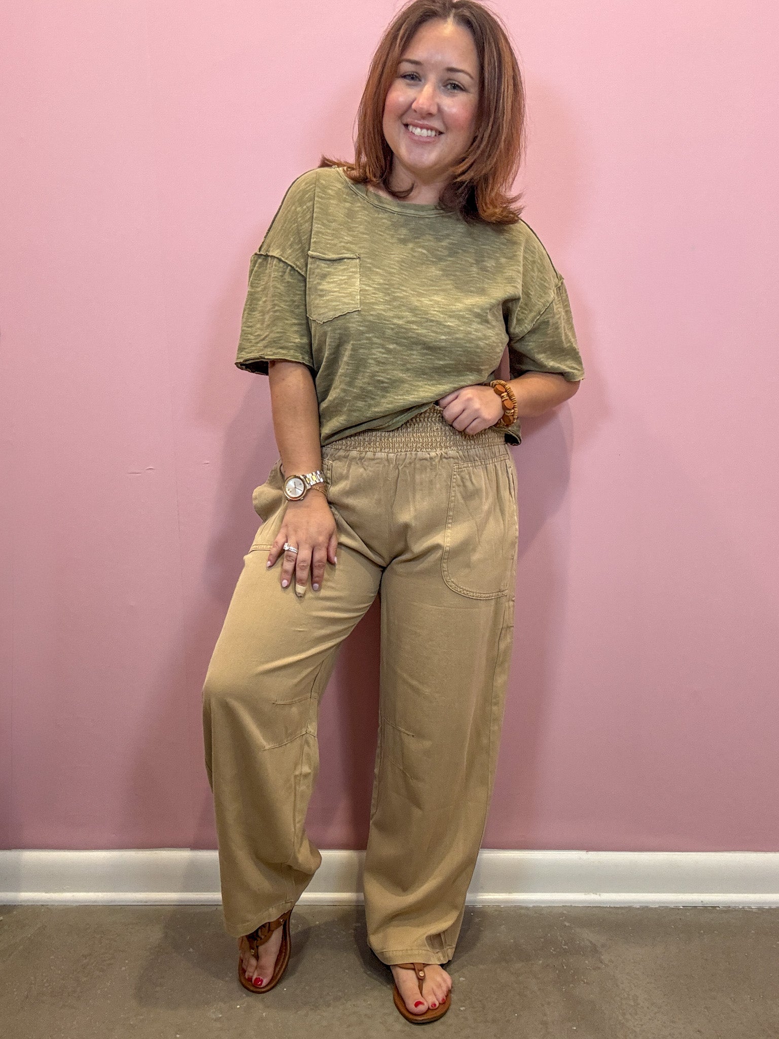 A woman modeling a two-piece outfit consisting of a short-sleeve olive green top and matching beige wide-leg pants, standing against a pink wall.
