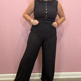 A woman modeling a black sleeveless jumpsuit with a button-down front and wide-leg pants, standing against a pink wall.