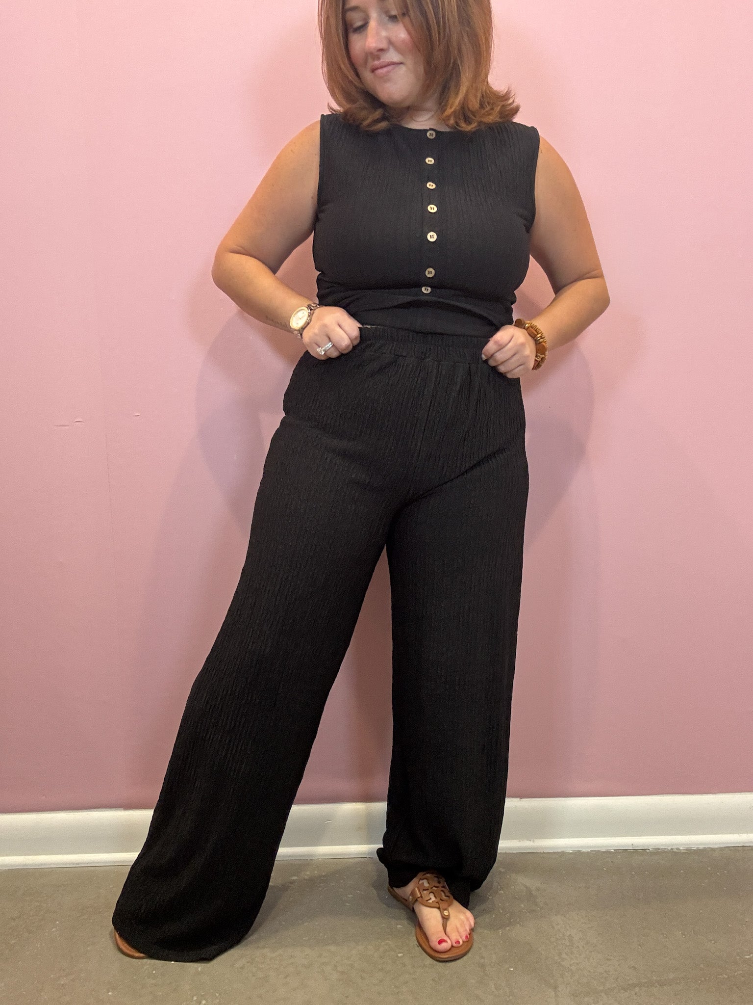 A woman modeling a black sleeveless jumpsuit with a button-down front and wide-leg pants, standing against a pink wall.