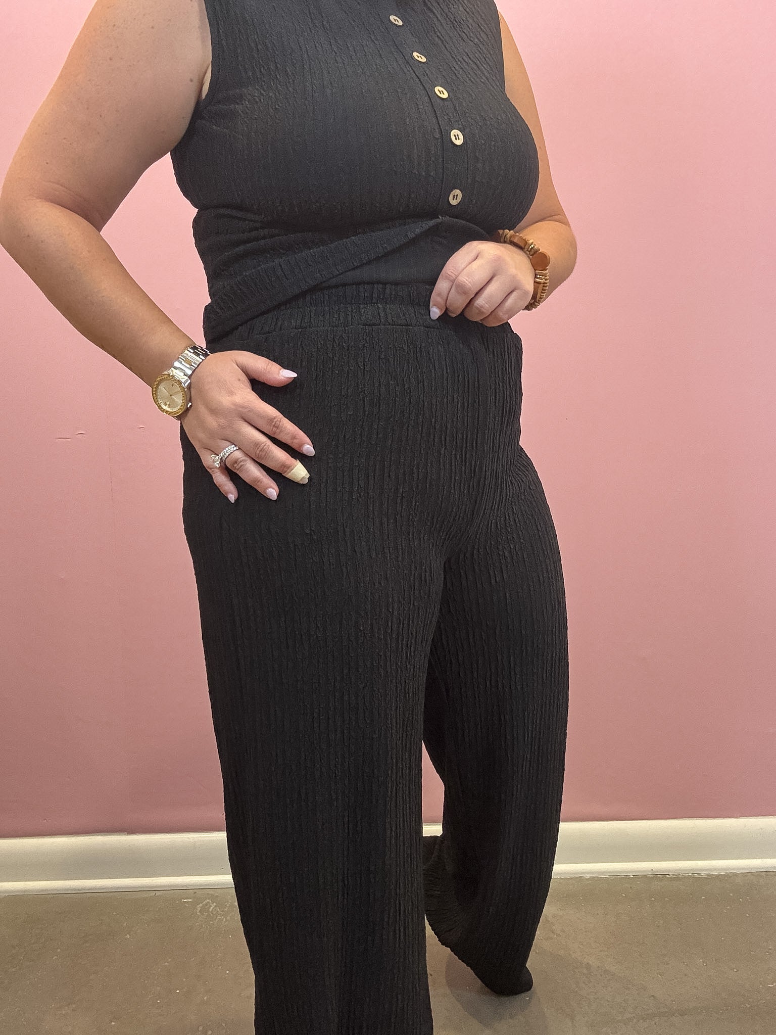 A person wearing a sleeveless black ribbed knit top with gold buttons and matching wide-leg black ribbed pants, standing against a pink wall.