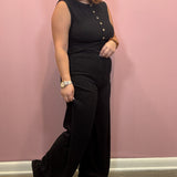 A woman modeling a black sleeveless jumpsuit with gold buttons down the front, paired with brown leather sandals, standing against a pink wall.